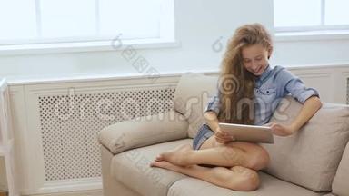 漂亮的少女在沙发上用平板电脑放松。 使用平板电脑和平板电脑在沙发上放松的一个可爱女孩的照片